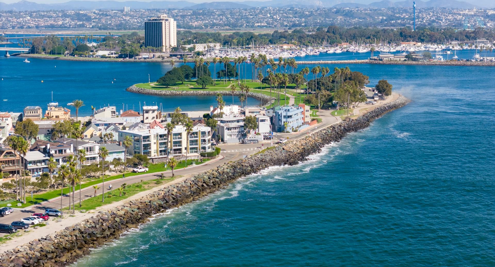 A sunny coastal town with a marina, palm-lined seawall and buildings beside turquoise water and distant mountains.
