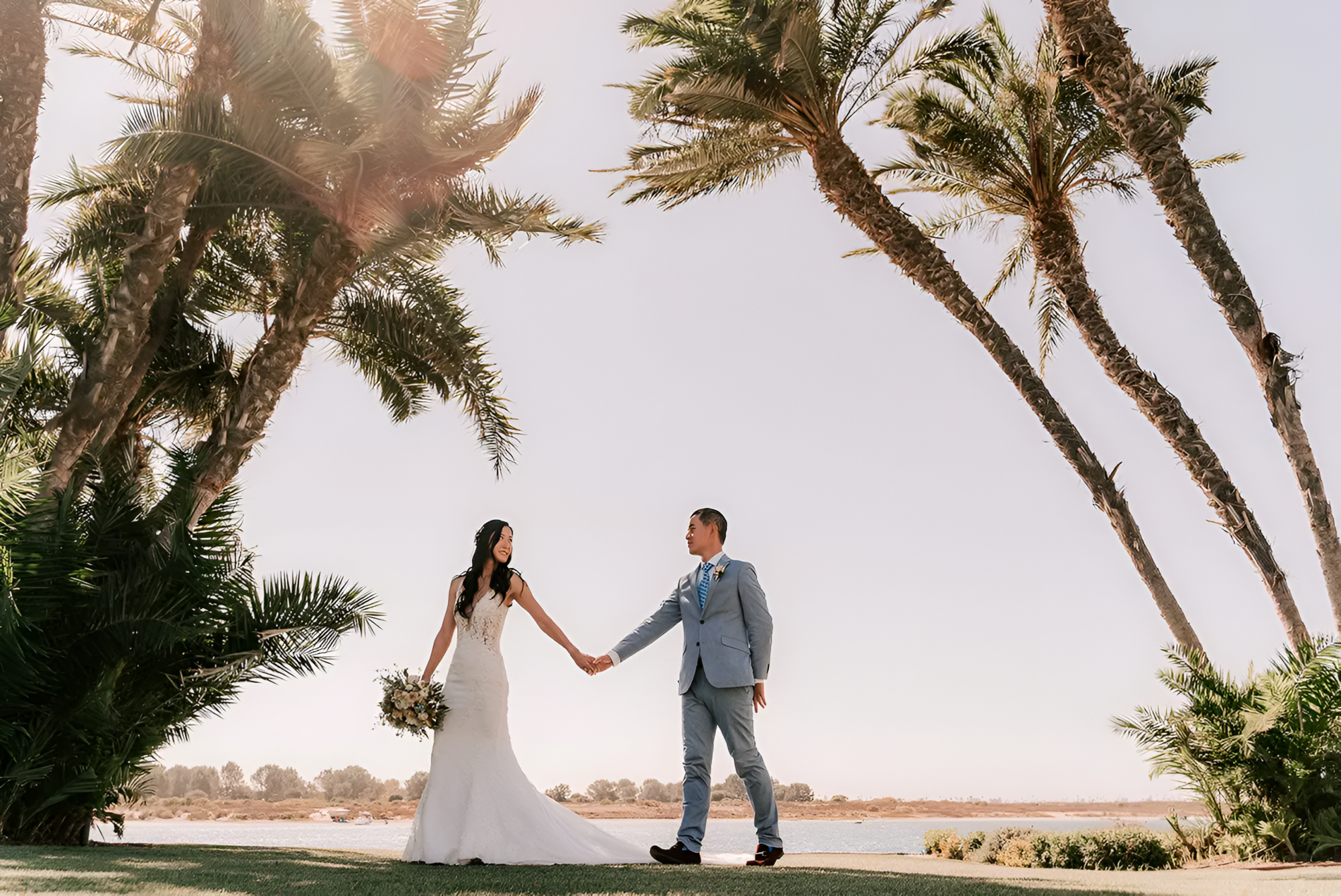 A bride and groom hold hands on a sandy overlook, surrounded by tall palm trees and a sunset-lit sky, celebrating their wedding vows.