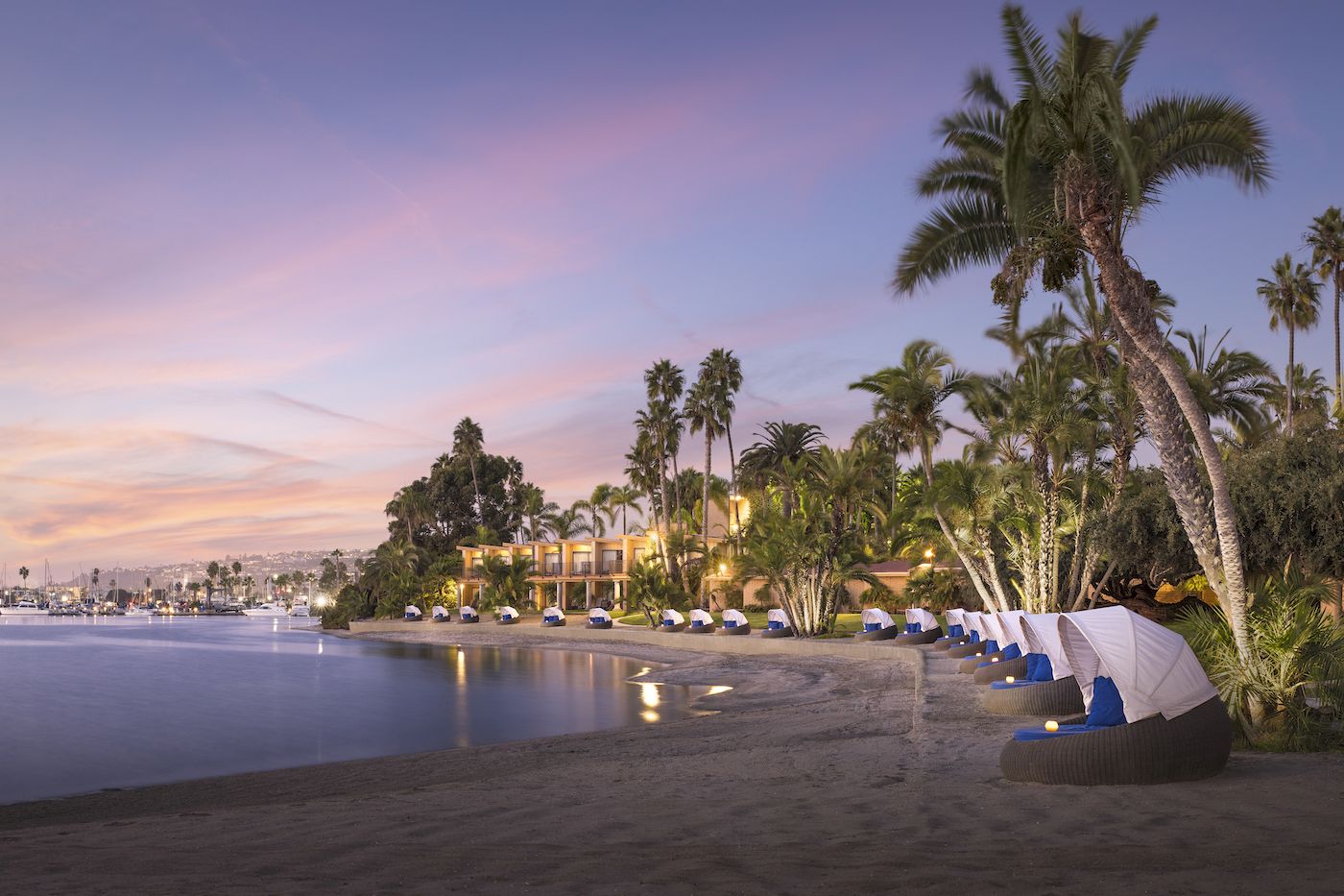 Palm-lined waterfront resort at dusk with white cabanas and glowing lights along a calm marina.