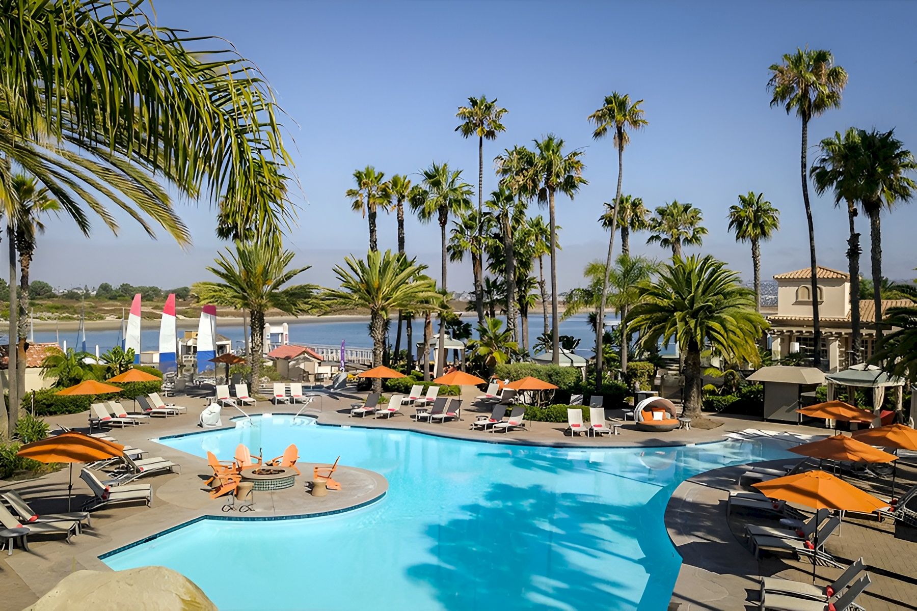 A sunny resort pool area with palm trees, lounge chairs, umbrellas, and people swimming and relaxing by the water, overlooking a calm view.