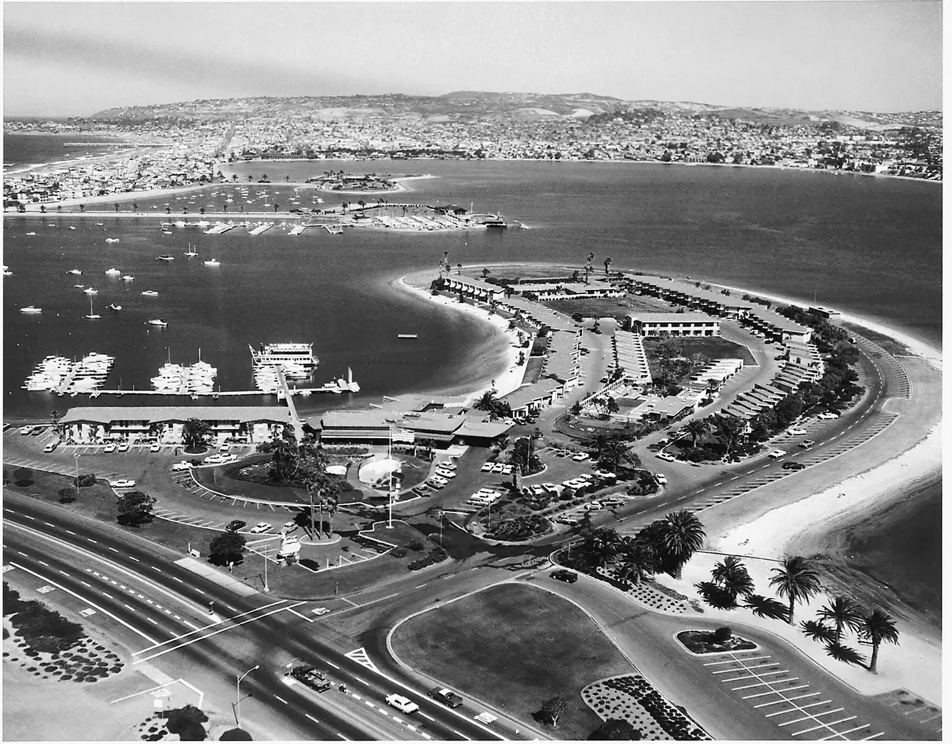 Aerial view of a curved harbor with a breakwater, marina docks, boats, palm trees, and a winding road along the waterfront.