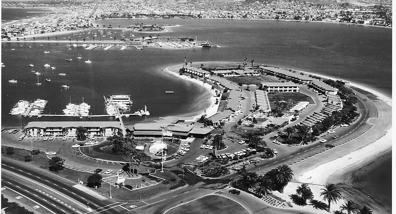 Aerial view of a curved harbor with a breakwater, marina docks, boats, palm trees, and a winding road along the waterfront.