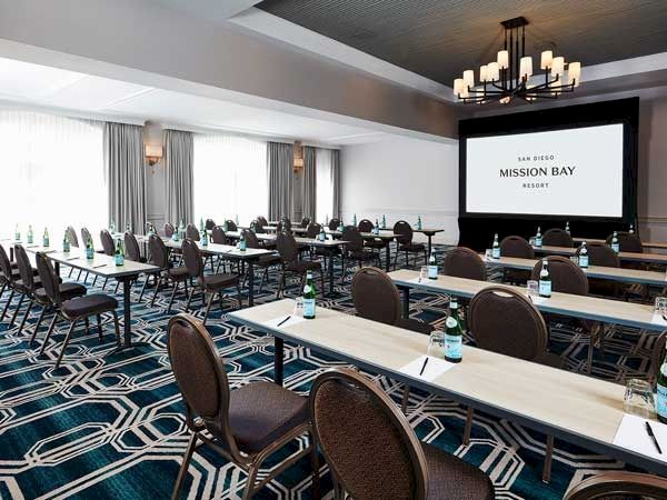 A conference room setup with long desks, brown chairs, water bottles, and a projector screen at the front displaying &ldquo;Mission Bay...&rdquo; conference style.