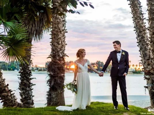 A bride in a white gown and a groom in a dark suit hold hands by palm trees on a lakeside at sunset.
