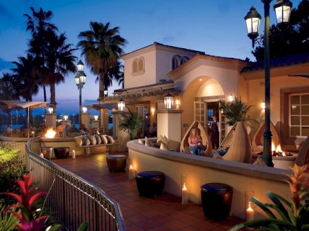 A luxurious seaside restaurant terrace at dusk with warm lights, palm trees, and guests lounging by the oceanfront, elegant architecture, and hanging lanterns.