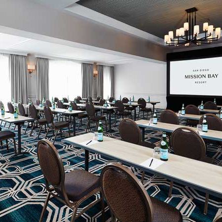 A conference room set up for a meeting with rows of desks, chairs, water bottles, and a large screen displaying &ldquo;Mission Bay Resort&rdquo; at the front.