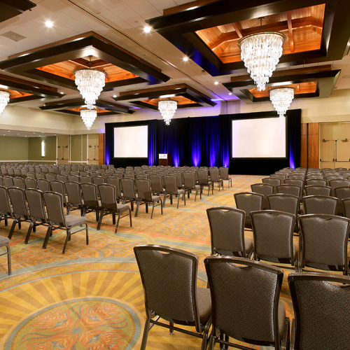 A large conference hall with rows of chairs, chandeliers, and a stage with blue-lit backdrop for presentations, ready for an event.