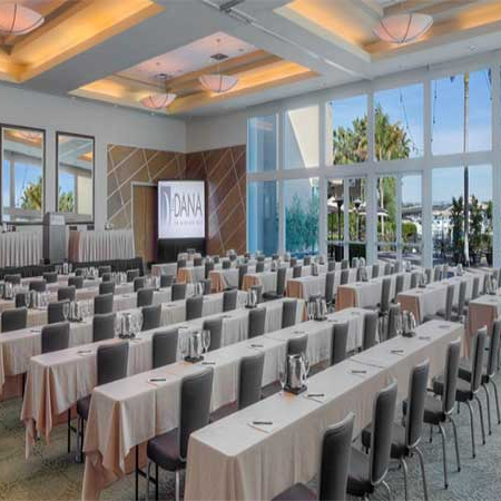 A spacious conference room set up for a seminar with rows of tables, pink tablecloths, gray chairs, and a large screen at the front, bright natural light from the windows, and a modern ceiling design.