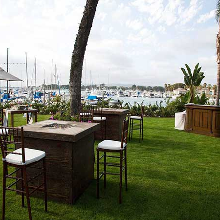 A stylish outdoor seating area on a grassy lawn near a marina, with high-top tables, chairs, palm trees, and water with boats in the background.