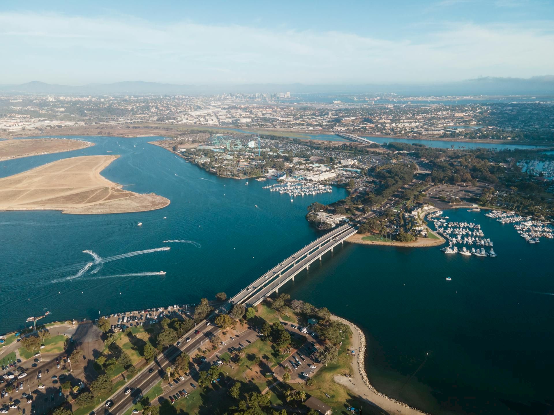 Aerial view of a wide river with a long bridge, marina, boats, and a coastal town under a clear sky.