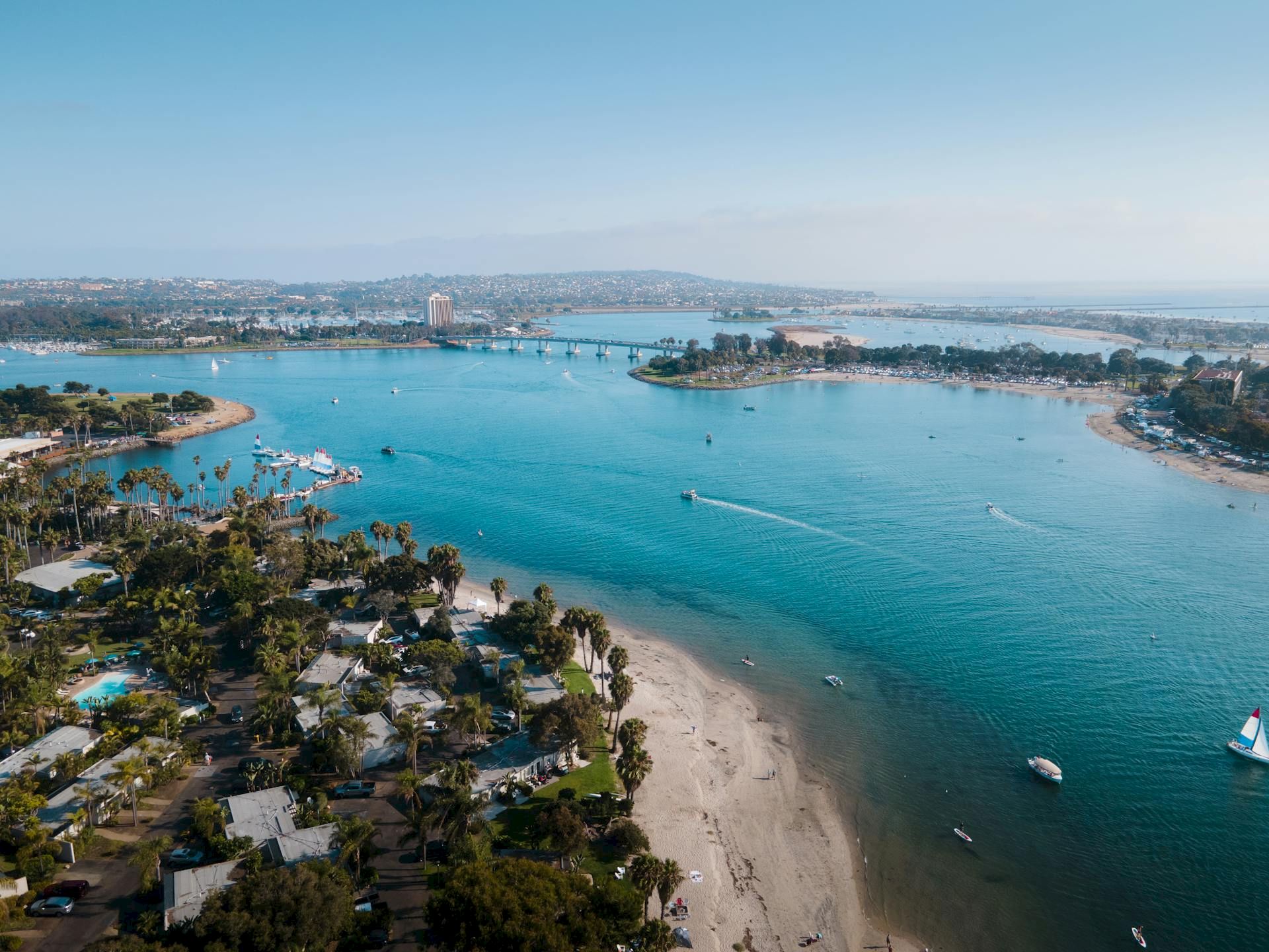 An aerial view of a sunny coastline with a harbor, long bridge, yachts and boats, and palm-lined beaches.