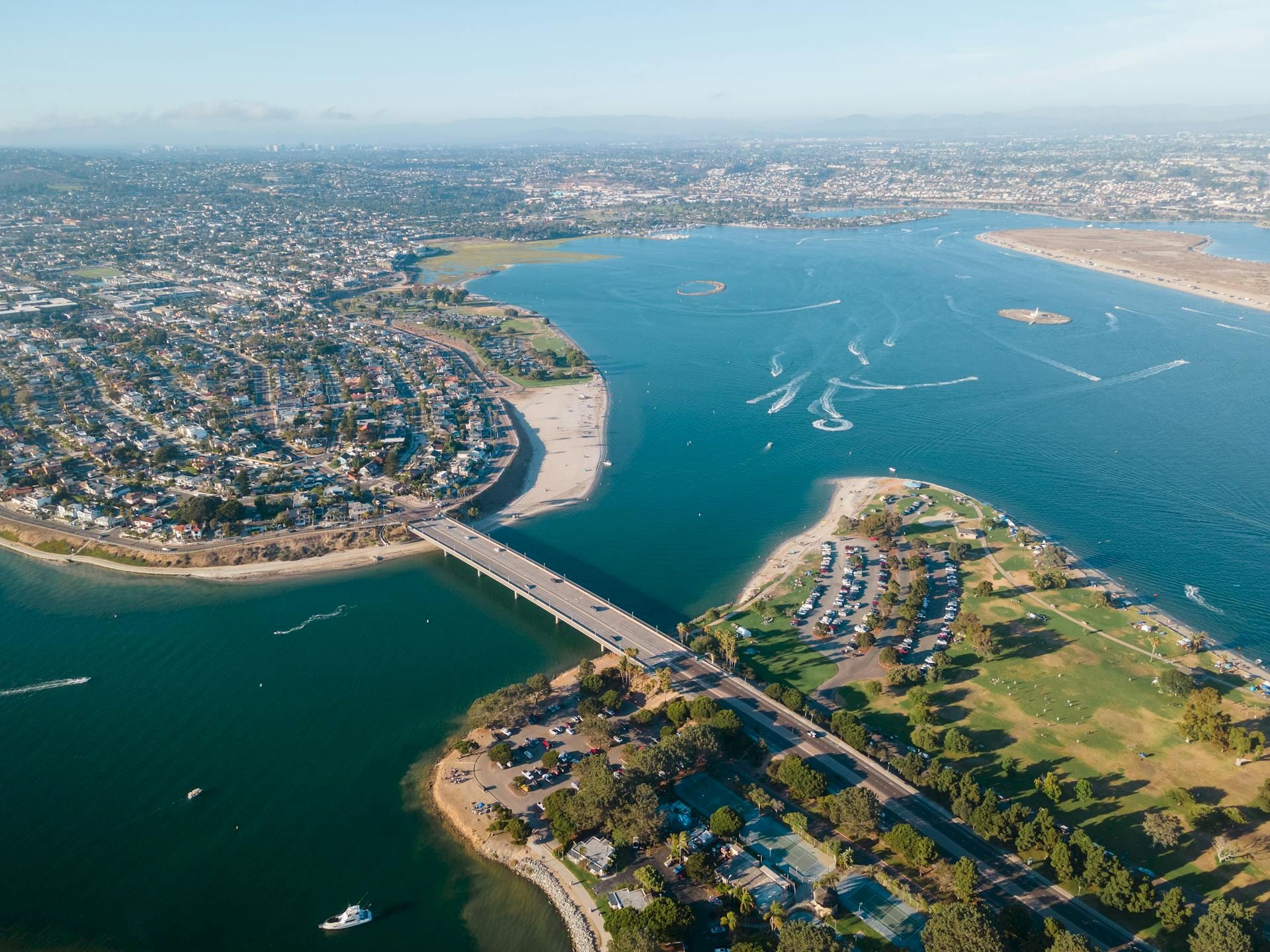Aerial view of a coastal city with a river or bay, a bridge crossing the water, dense housing on both sides, and boats in the water.