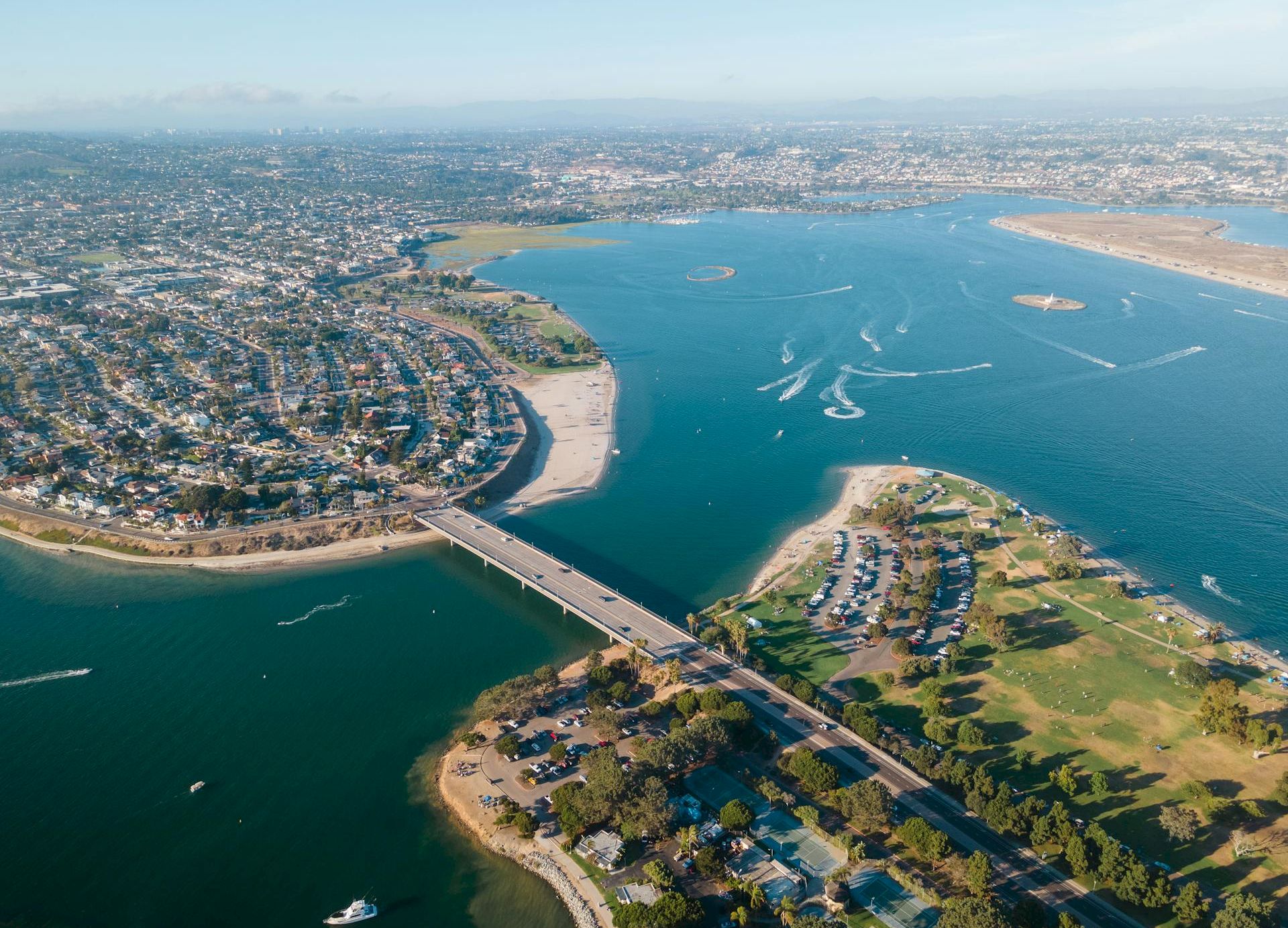 Aerial view of a coastal city with a river or bay, a bridge crossing the water, dense housing on both sides, and boats in the water.