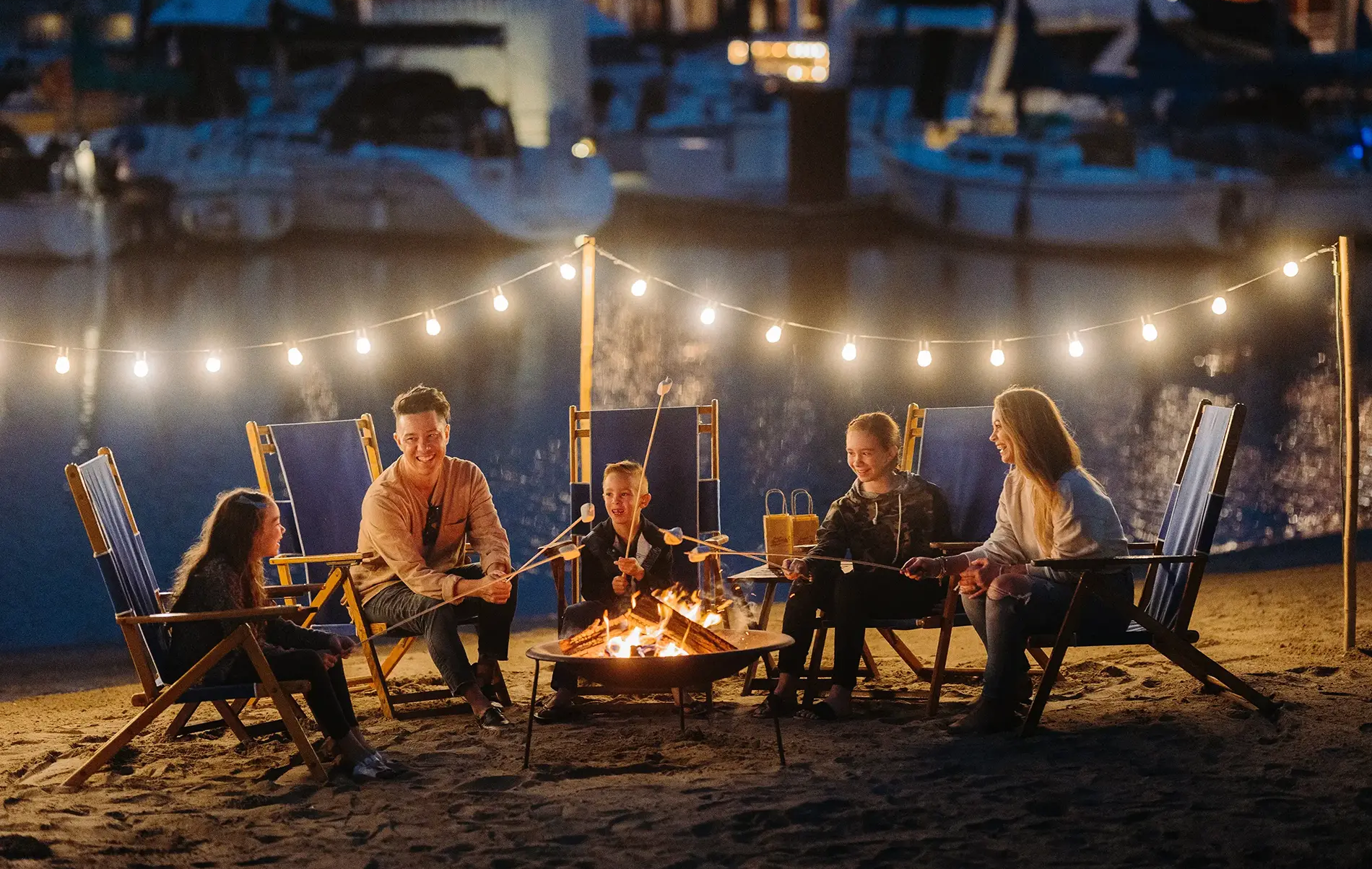 A group of friends sits around a glowing campfire on the beach, string lights overhead, boats in the harbor behind them, cozy night.