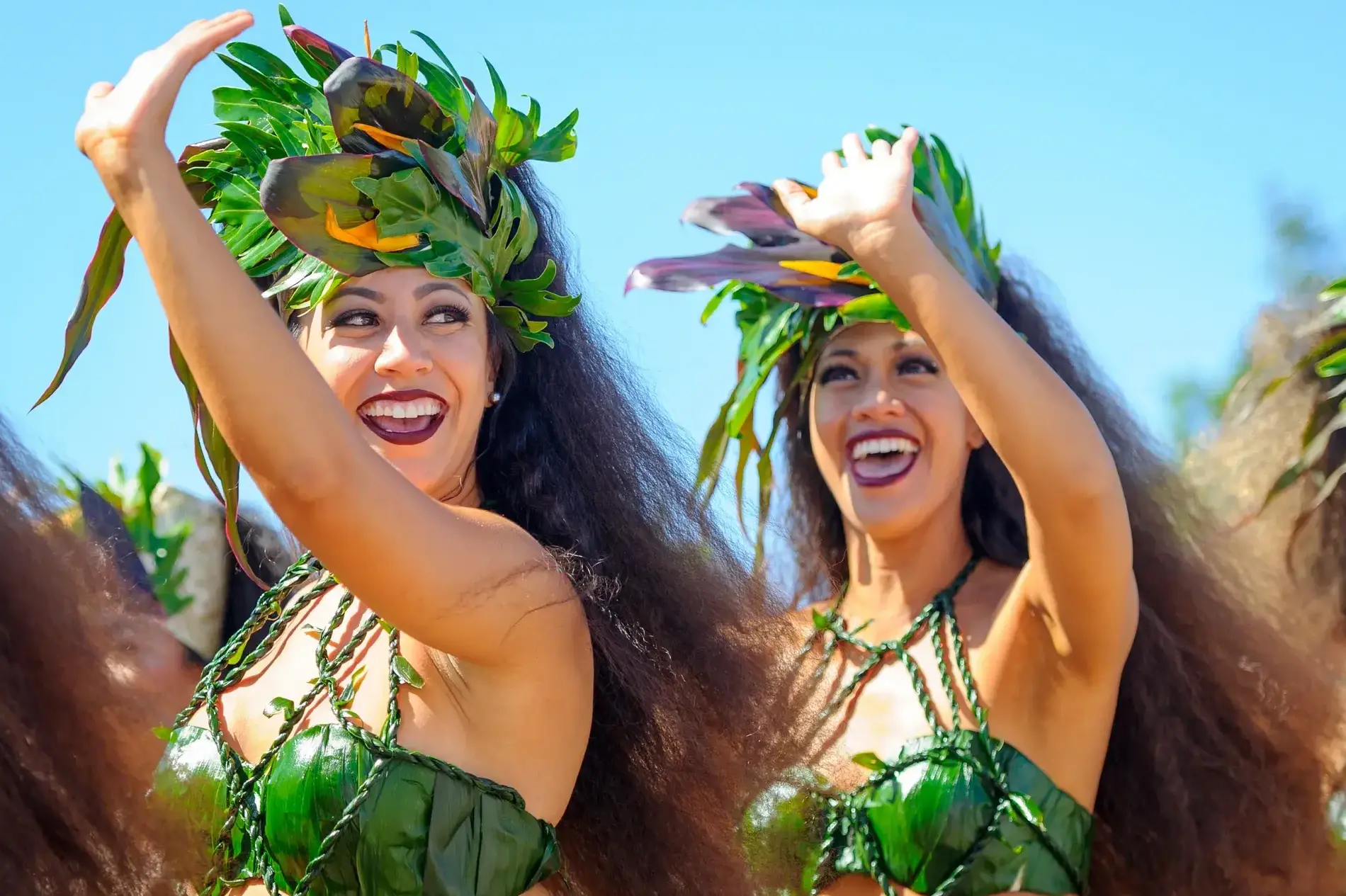 Two smiling women in green tropical outfits with leafy headdresses perform a dance, bright blue sky behind them.