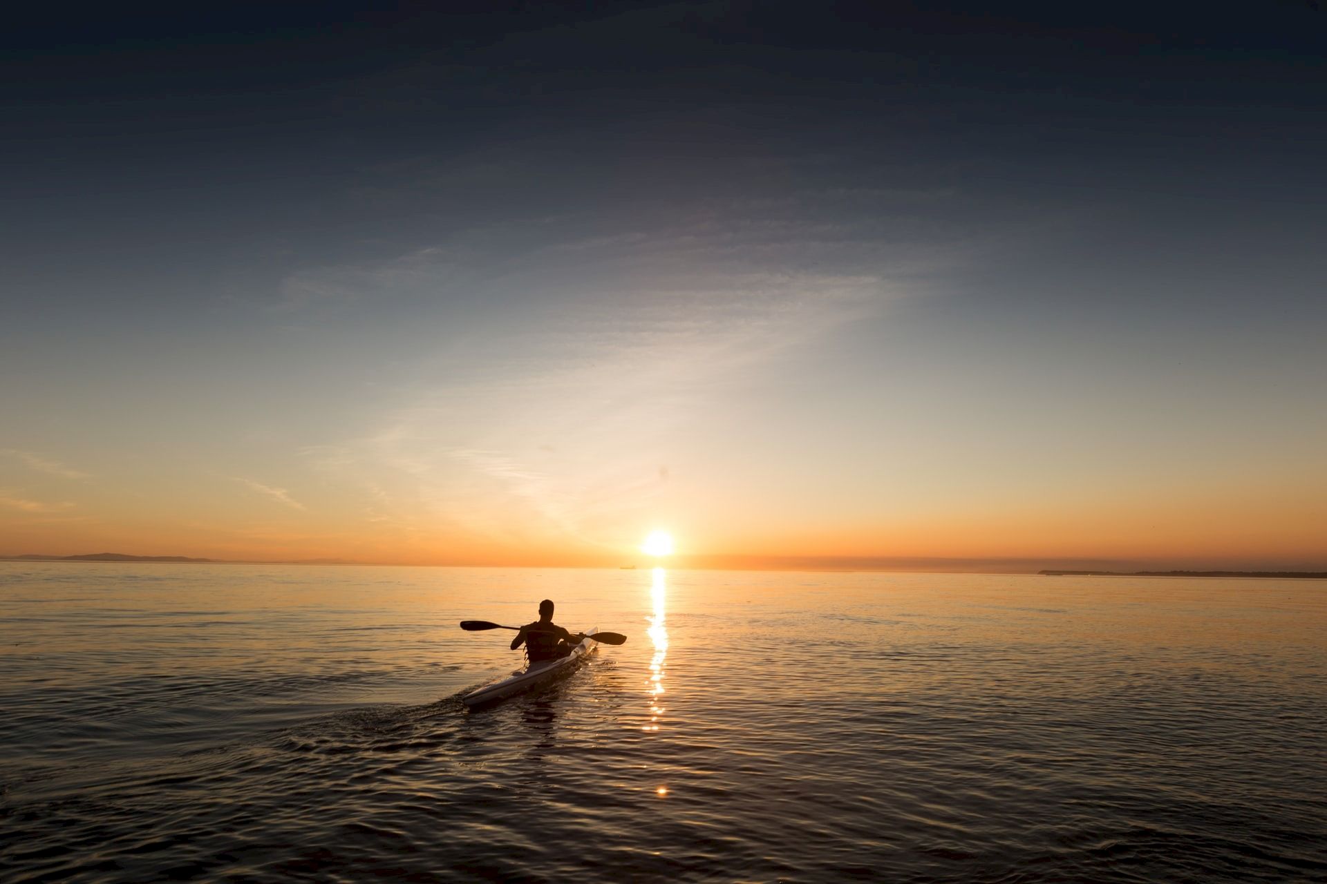 A lone kayaker paddles across calm water at sunset, with the sun&rsquo;s reflection shimmering on the sea.