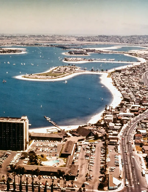 Aerial view of a coastal city with a curved shoreline, high-rise hotel near water, and a grid of colorful houses along a busy road.