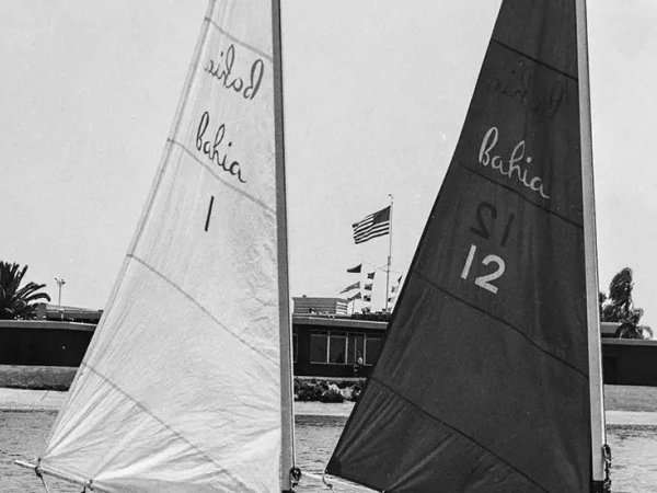 Two small sailboats race on a sunny beach with sails labeled &ldquo;Bahia,&rdquo; flags in background, and a calm shoreline behind.