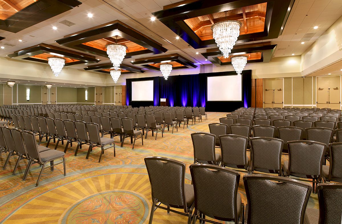 A large conference hall with many rows of chairs facing a stage and two screens, elegant chandeliers, and warm lighting.
