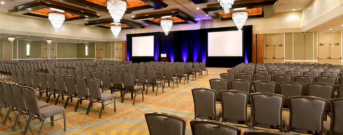 A large conference hall with many rows of chairs facing a stage and two screens, elegant chandeliers, and warm lighting.