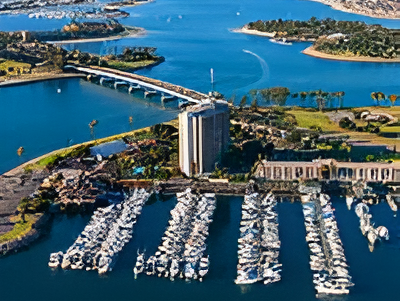 Aerial view of a marina with numerous boat slips, a tall building on the waterfront, a curved bridge, and surrounding coastline.