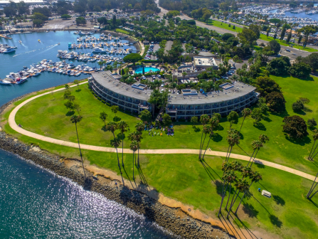 Aerial view of a curved, circular building by the water, surrounded by green lawns, palm trees, a marina, and winding paths along the coastline.