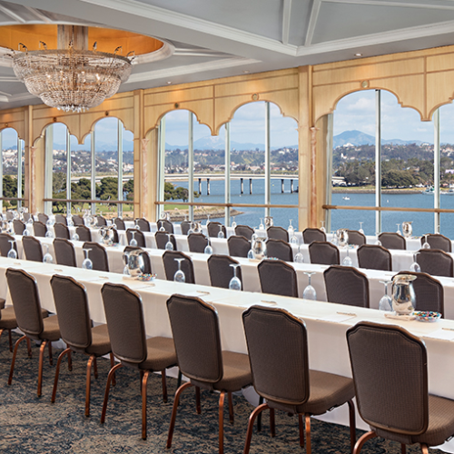 A sunlit banquet room with long white-table setup, cushioned chairs, chandeliers, and large bay windows overlooking a scenic waterfront.