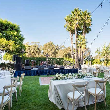 Outdoor wedding setup with round and rectangular tables, white linens, floral centerpieces, string lights, a stage, and palm trees on a sunny lawn.
