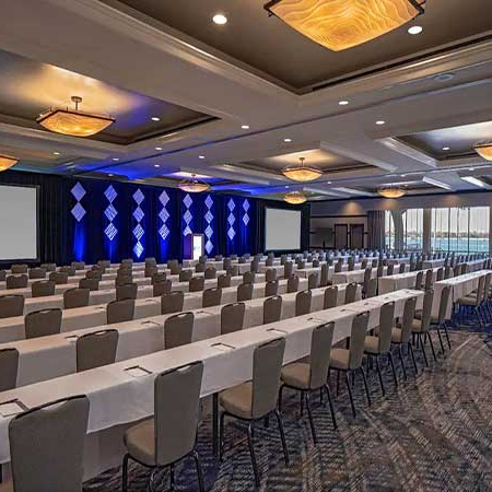 A large conference room set up with long rows of beige chairs and white tablecloths, stage with blue lighting, and two projection screens, ready for a seminar.