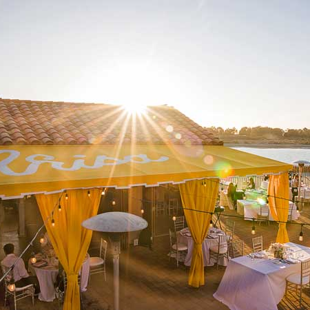 A sunlit outdoor party with a yellow canopy, string lights, tables, and guests mingling near the waterfront at sunset.