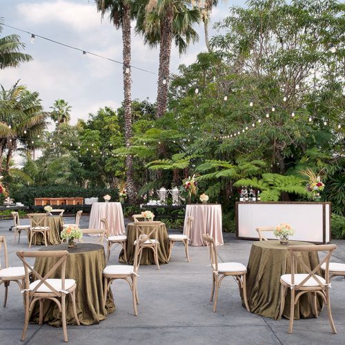 Outdoor event setup with round and rectangular tables, neutral tablecloths, wooden chairs, string lights, and lush green trees in a garden.