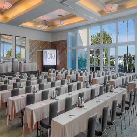 A modern conference room set for a seminar, with long rows of desks and chairs, a stage with a screen, bright natural light, and ceiling lights.