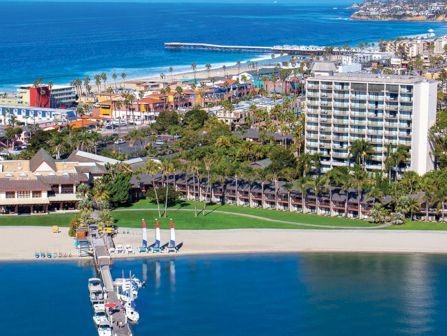 Sunny beach resort with white buildings, palm trees, a wooden pier, and calm turquoise water stretching to a distant pier and skyline.