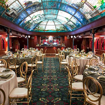 A luxurious banquet hall with round tables, elegant chairs, floral centerpieces, and a colorful glass ceiling, set for a formal event.