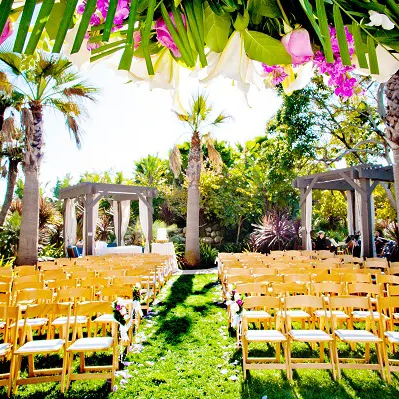 A sunny outdoor wedding setup with rows of wooden chairs facing an aisle, under palm trees and decorated arches, vibrant greenery and colorful flowers.