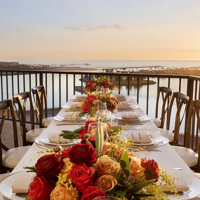 A long balcony table set for a sunset dinner with white plates, floral centerpieces of red and peach roses, and a scenic water view in the distance, ending gracefully.