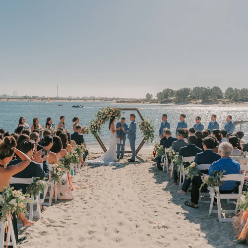 A beach wedding ceremony: couple in formalwear under a floral arch, guests seated along a sandy aisle by the water.