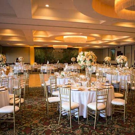 A formal banquet setup with round tables, white tablecloths, elegant floral centerpieces, and gold-toned chairs in a well-lit event hall.