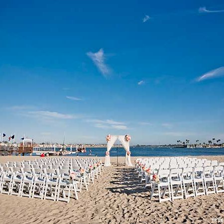 A beach setup with rows of white chairs facing an archway by the shore, perfect for a wedding ceremony, under a clear blue sky.