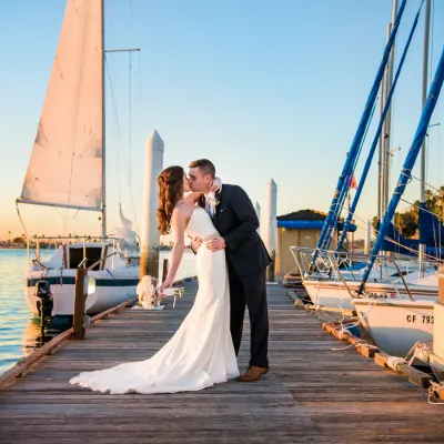 A bride and groom kiss on a wooden pier by calm water with sailboats, sunset glow, and clear blue sky, capturing a romantic wedding moment.