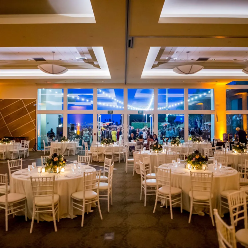 A stylish banquet hall with round tables, white chairs, floral centerpieces, warm lighting, and large windows showcasing a cityscape, ready for a reception.