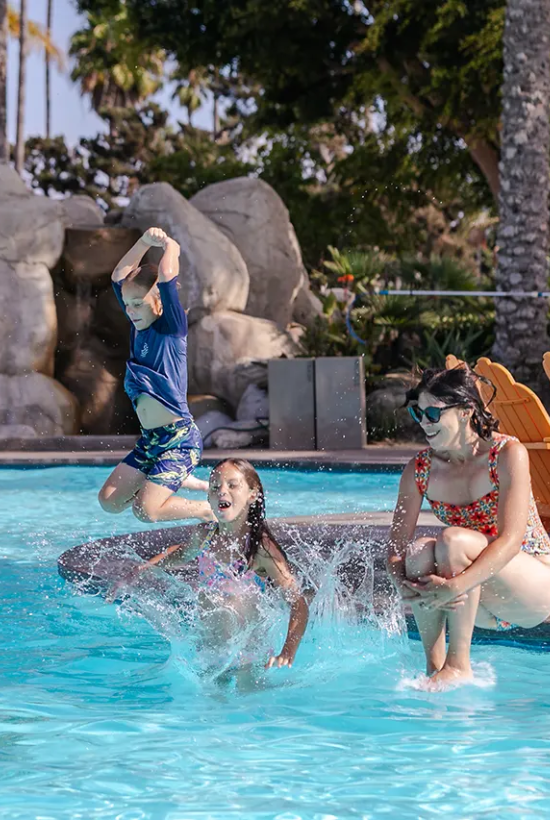 People enjoy a pool: two kids splash in, a man jumps from rocks, and two women sit by the water near orange lounge chairs.