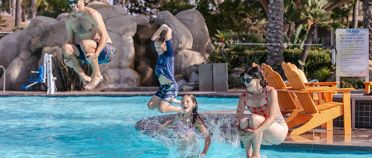 Two people jump into a sunny pool while two others watch from the edge; palm trees and rocks frame a resort scene with bright chairs in the background.