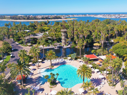 Aerial view of a tropical resort with a large irregular blue swimming pool surrounded by palm trees, lounge chairs, walkways, and a lake or sea in the distance.