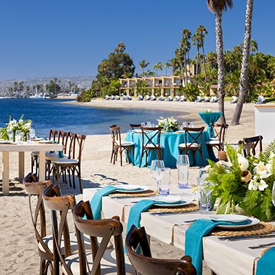 A beachside dining setup with white-clothed tables, turquoise accents, flower centerpieces, and wooden chairs along the sandy shore under palm trees.