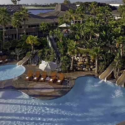 A luxury resort pool area with palm trees, lounge chairs, umbrellas, and curvy blue pools surrounded by lush greenery.