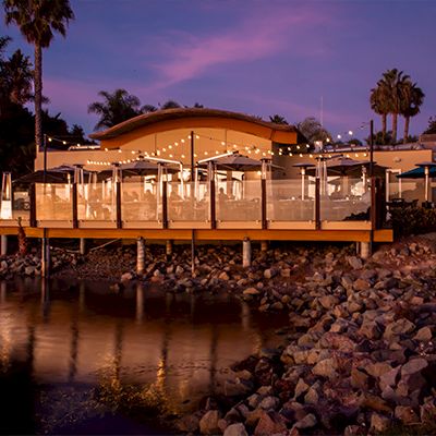 A waterfront restaurant with a wooden deck, patio seating, umbrellas, palm trees, and calm water at dusk.