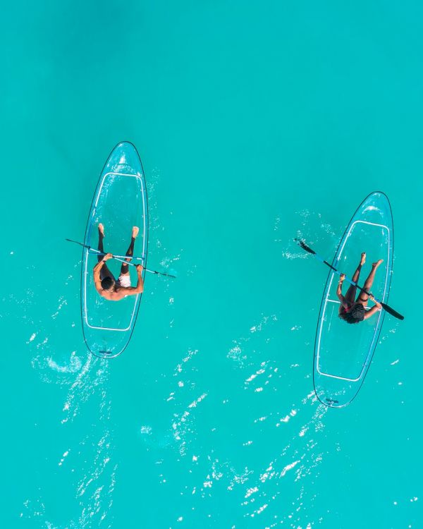 Two people stand on separate transparent surfboards riding turquoise water, paddling apart in calm, crystal-clear sea, enjoying sunny waves.