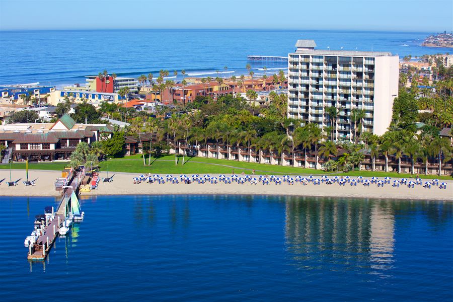 A sunny beach resort with a long sandy shore, turquoise water, a pier, palm trees, and a tall white hotel overlooking the ocean, busy with people.