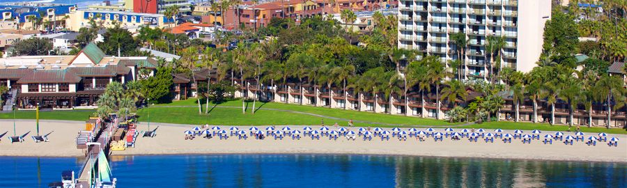 A sunny beach resort with a long sandy shore, turquoise water, a pier, palm trees, and a tall white hotel overlooking the ocean, busy with people.